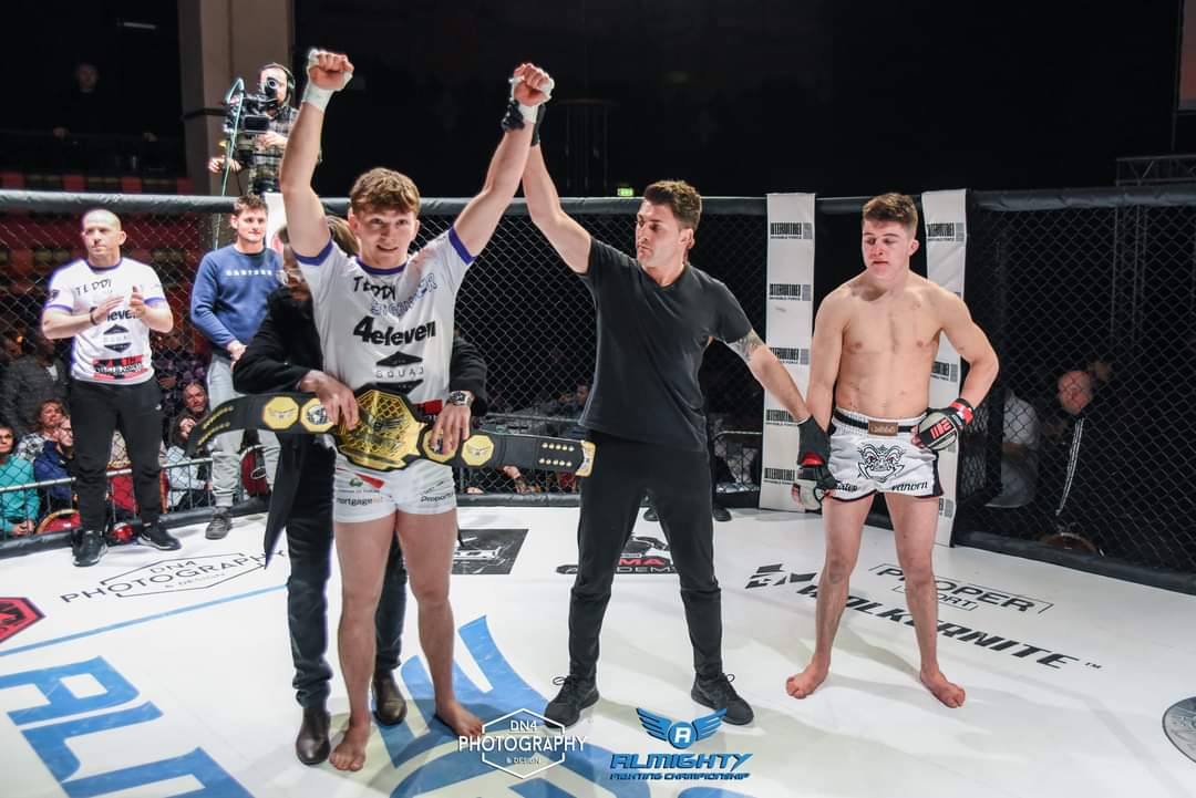 Fighter in white 4 Eleven kit having his hand raised as champion with the belt beside him